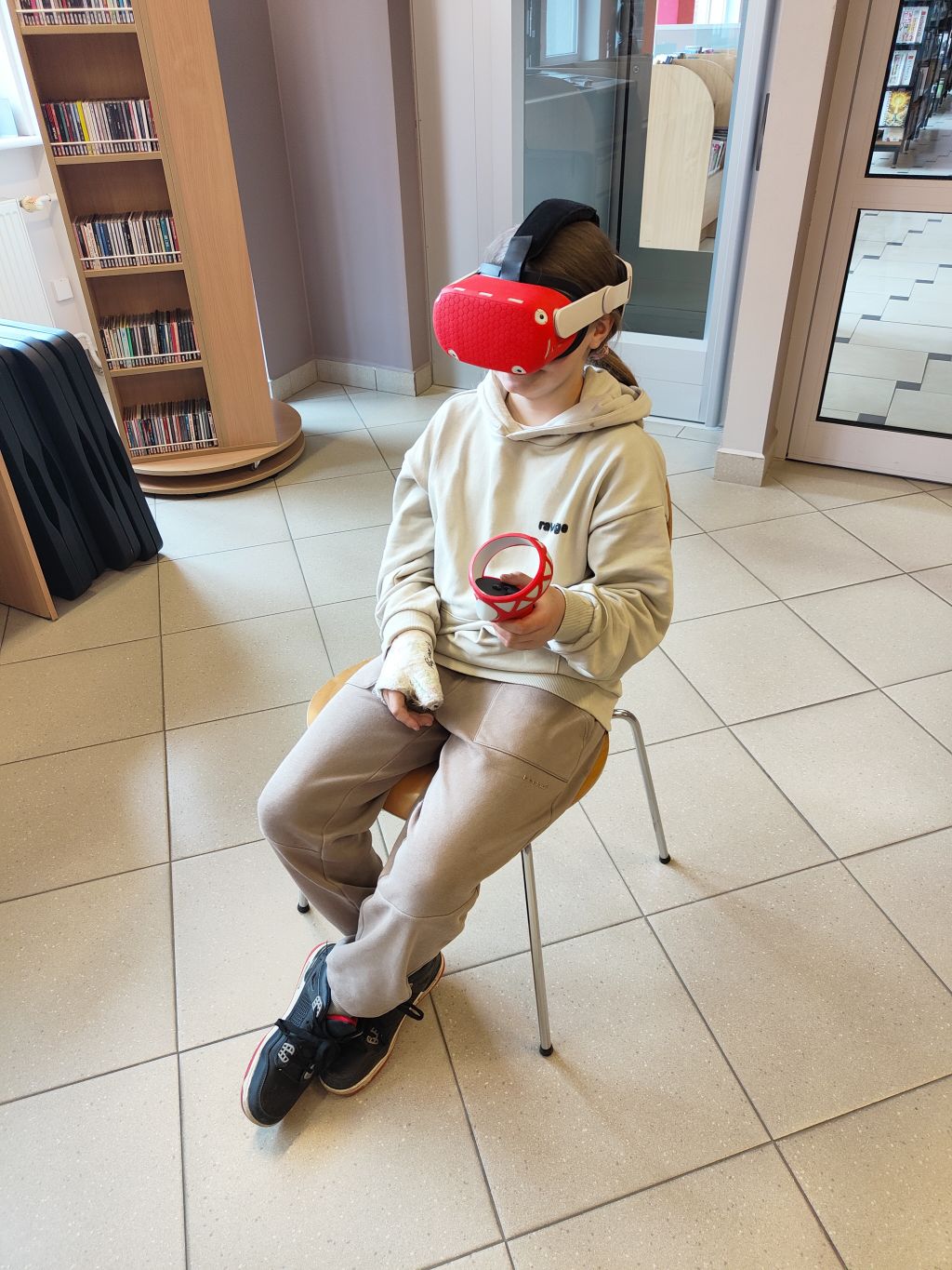 Dziecko siedzące na krześle, ubrane w beżową bluzę i spodnie dresowe, ma na twarzy gogle VR z czerwoną osłoną. Trzyma kontroler z czerwoną obwódką w jednej ręce; druga ręka jest owinięta bandażem. Widok z góry.