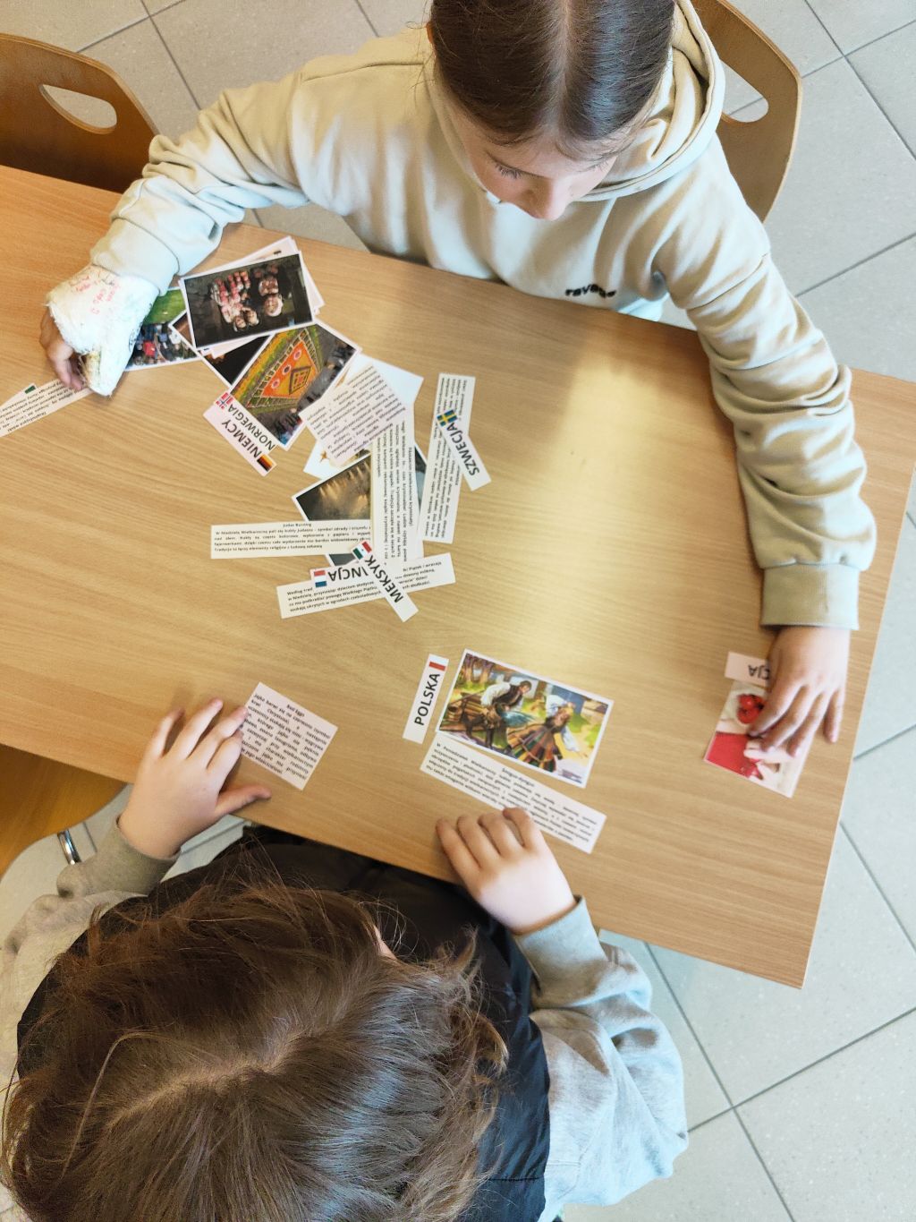 Ujęcie z góry przedstawiające dwoje dzieci przy stole układających karty z nazwami państw (POLSKA, SZWECJA, MEKSYK, NIEMCY) oraz odpowiadające im zdjęcia kulturowe. Jedna ręka dziecka jest zabandażowana.