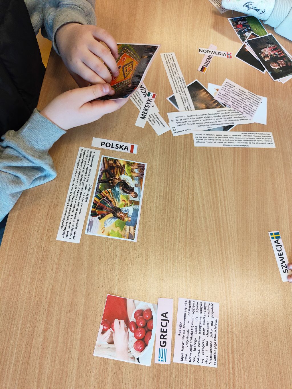 Zbliżenie na stół, przy którym dwoje dzieci układa i przyporządkowuje zdjęcia do napisów z nazwami państw (POLSKA, MEKSYK, NORWEGIA, NIEMCY, GRECJA). Widać ręce dzieci, w tym jedną zabandażowaną.
