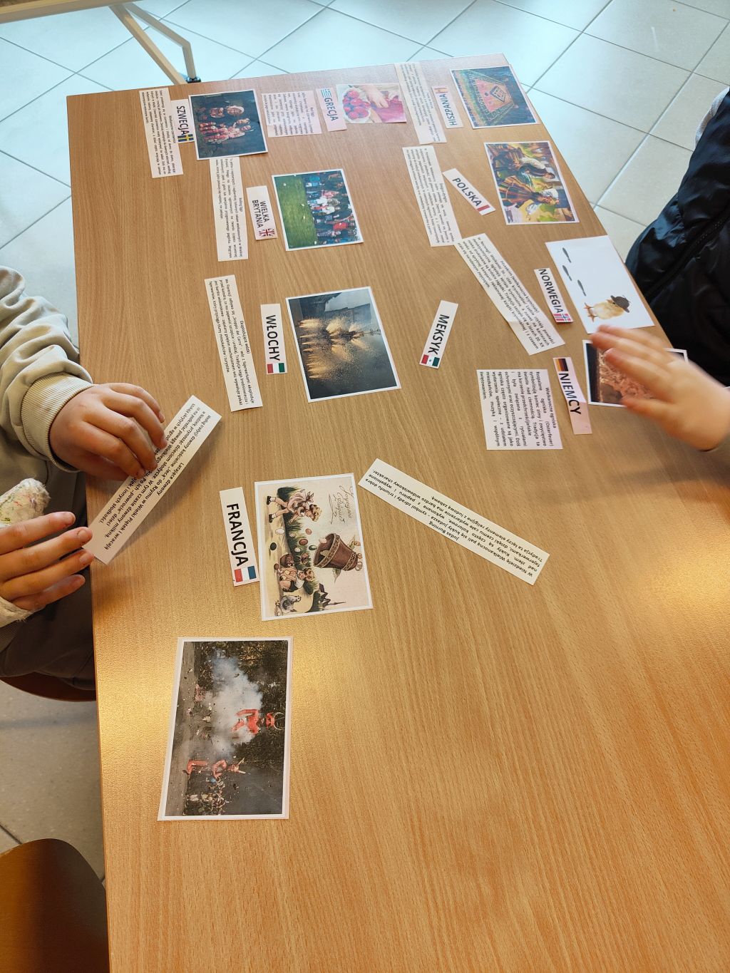 Widok z góry na drewniany blatu stołu, na którym rozłożone są kartki ze zdjęciami i napisami z nazwami krajów po polsku (np. WŁOCHY, FRANCJA, NORWEGIA). Widoczne ręce dwóch osób przesuwających materiały.