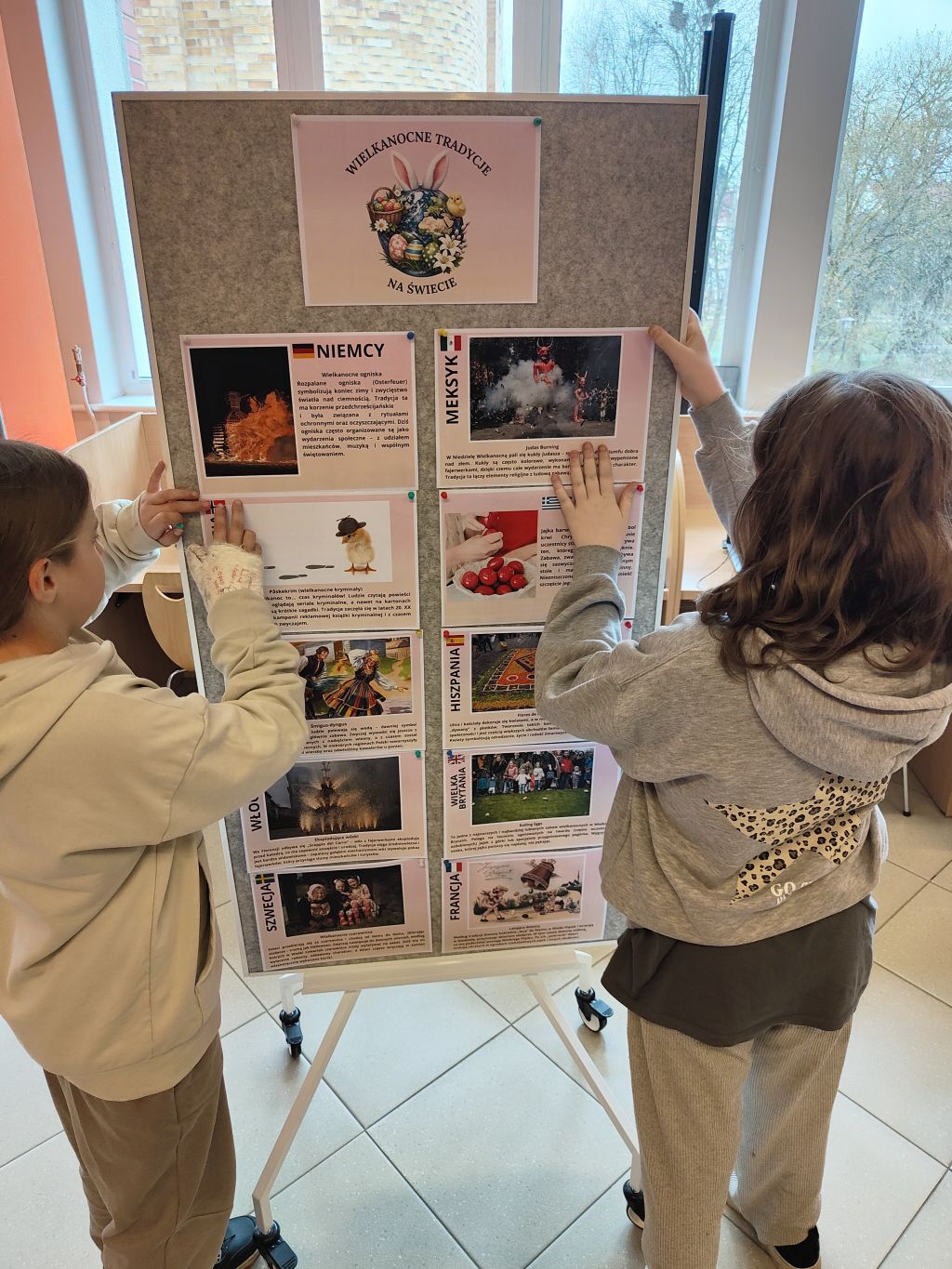 Dwoje dzieci przypina kartki do mobilnej tablicy korkowej na kółkach. Tablica ma nagłówek 