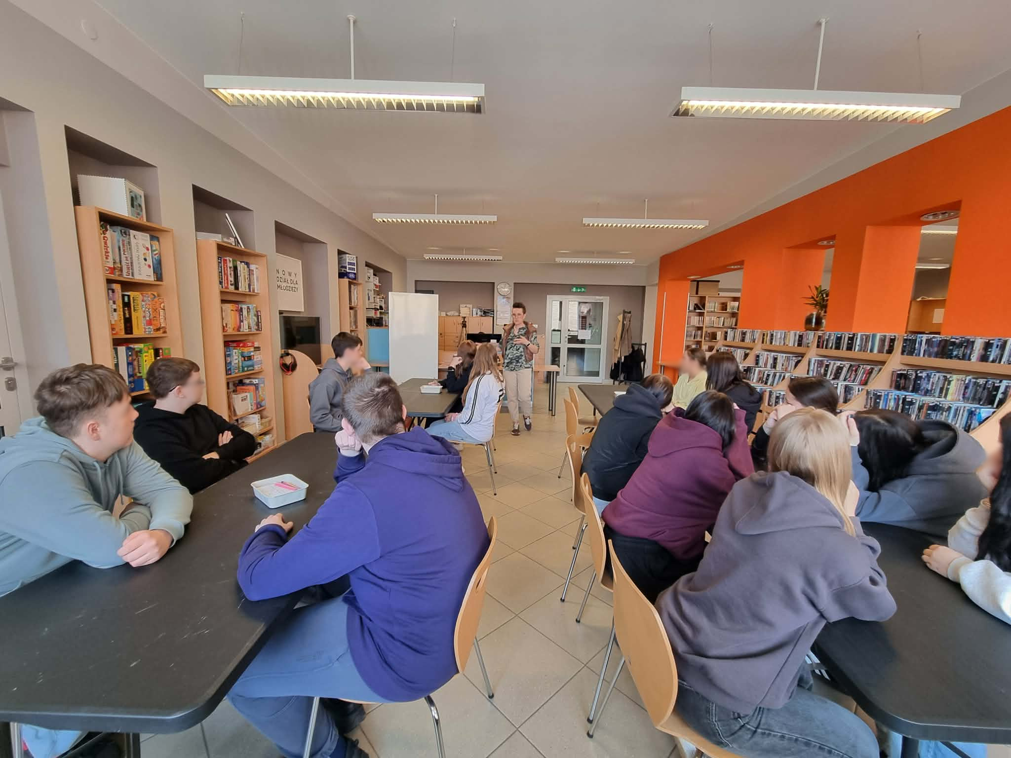 Prowadząca omawia temat zajęć, a młodzież siedzi przy stołach w przestrzeni bibliotecznej.