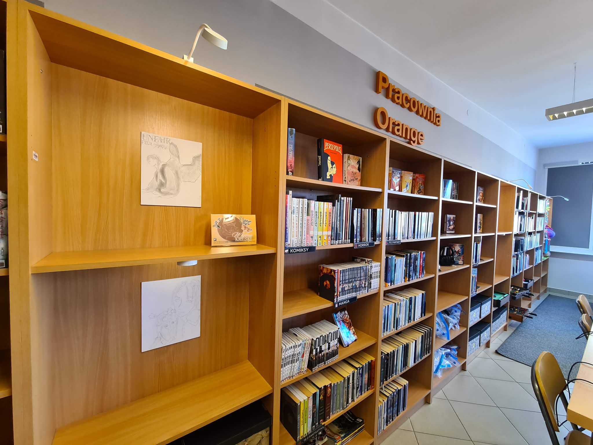 Widok regałów bibliotecznych w Pracowni Orange z wyeksponowanymi rysunkami i tabliczką wystawy „W rytmie kreski