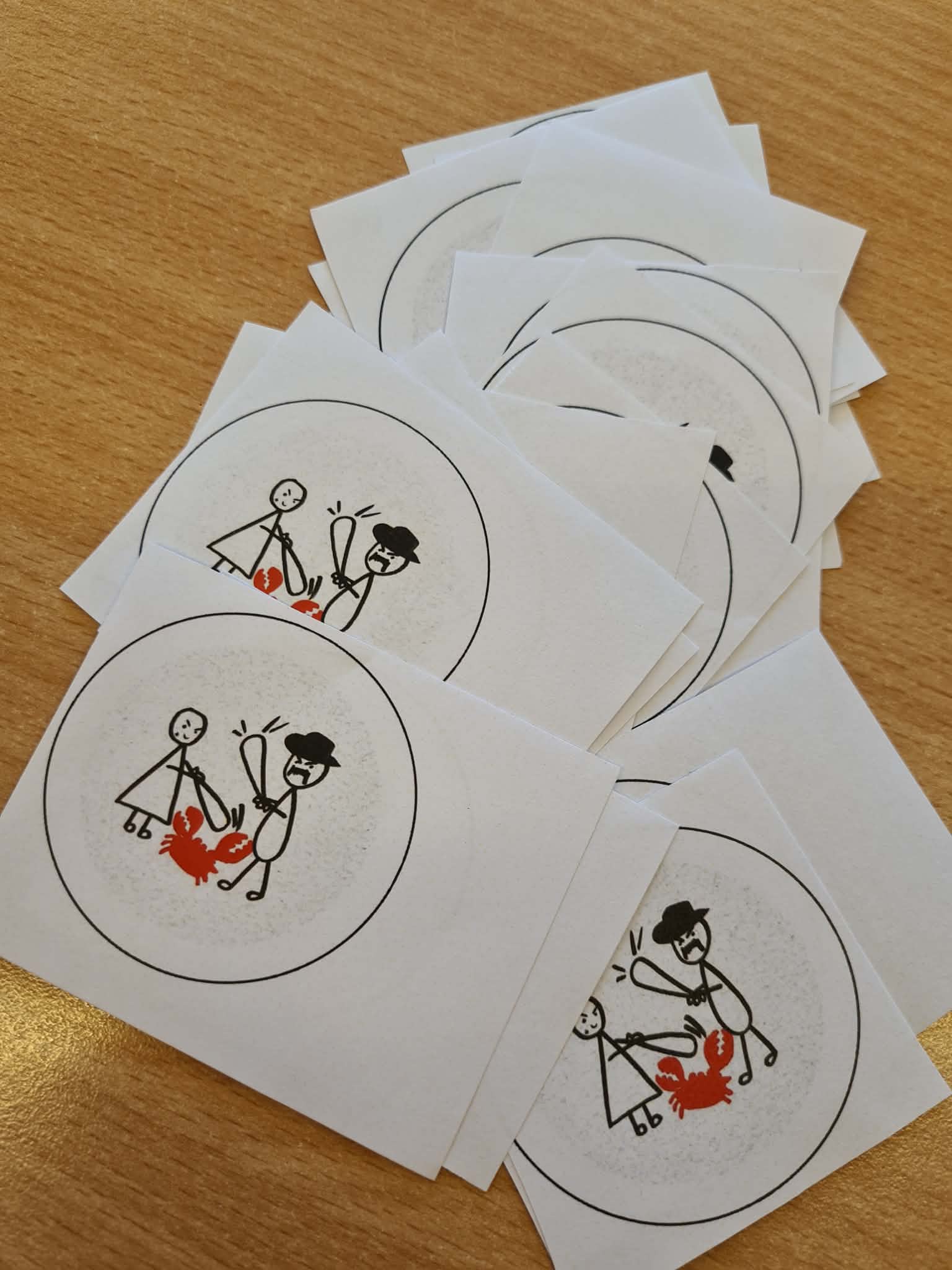 Stos wyciętych papierowych grafik z rysunkiem postaci i czerwonego raka, przygotowanych do wykonania przypinek.