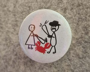 Gotowa biała przypinka z rysunkiem postaci i czerwonego raka przypięta do szarego materiału.