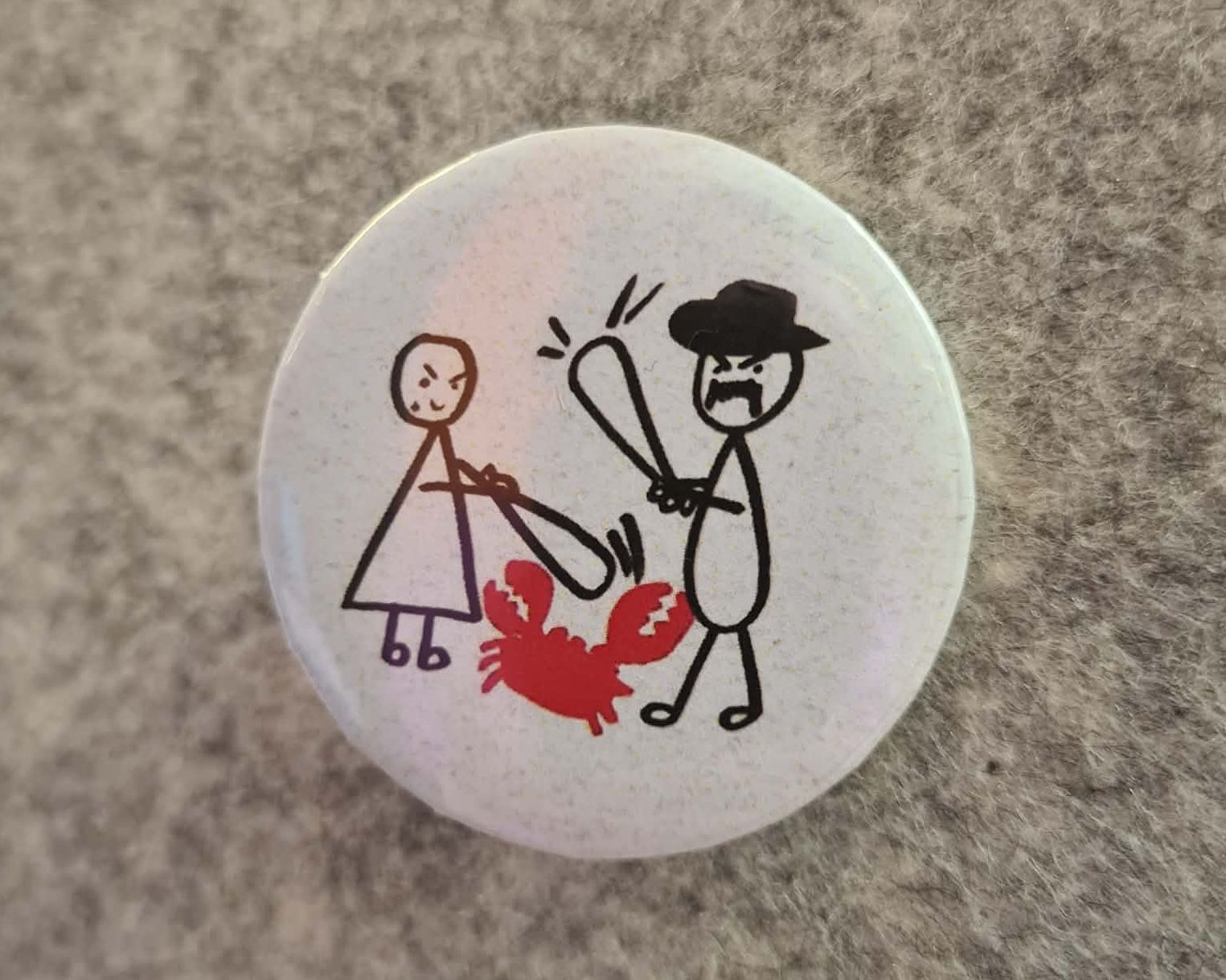 Gotowa biała przypinka z rysunkiem postaci i czerwonego raka przypięta do szarego materiału.