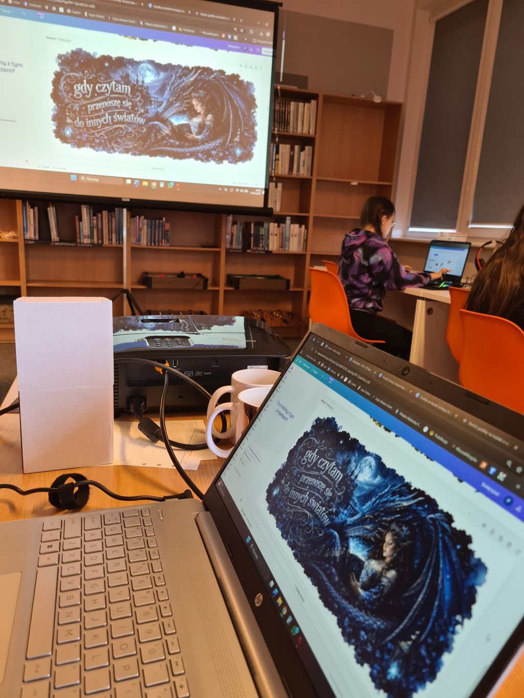 Sala warsztatowa w bibliotece. Na ekranie projektora i laptopie widoczny jest projekt grafiki na kubek z motywem fantasy i napisem „gdy czytam przenoszę się do innych światów”.