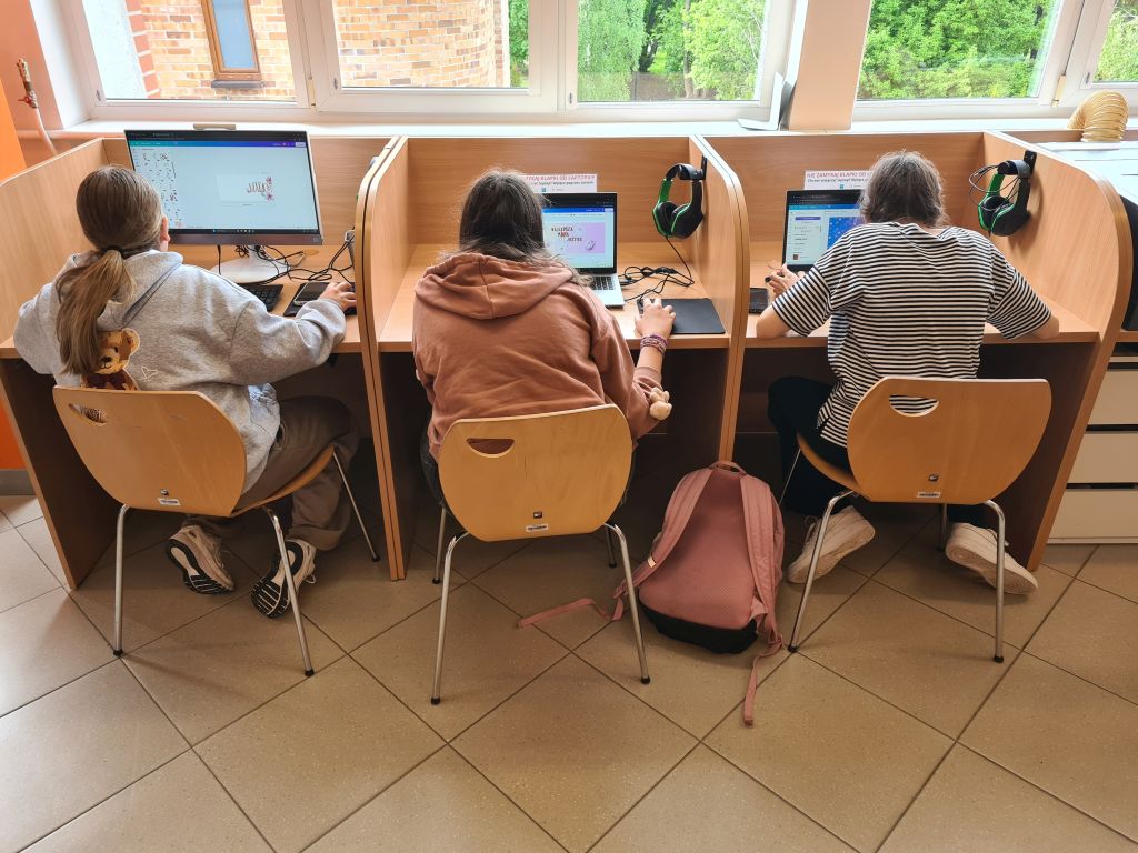 Trzy osoby siedzą tyłem przy osobnych biurkach z przegrodami, pracując na komputerach stacjonarnych i laptopach w jasnym pomieszczeniu z dużymi oknami.