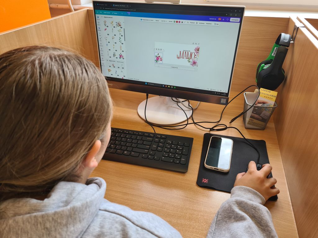 Widok zza ramienia na osobę projektującą grafikę z kwiatami i napisem „KOCHAM CIĘ MAMO” w programie komputerowym. Obok klawiatury leży smartfon.