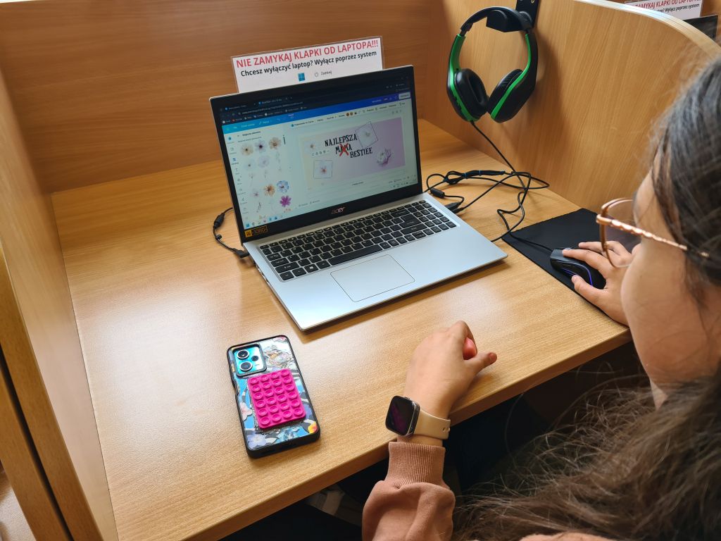 Osoba w okularach pracuje na laptopie Acer, używając myszki. Na ekranie widoczny jest projekt graficzny z napisem „NAJLEPSZA MAMA BESTIE”. Na biurku leży telefon w kolorowym etui z różową zabawką pop-it.