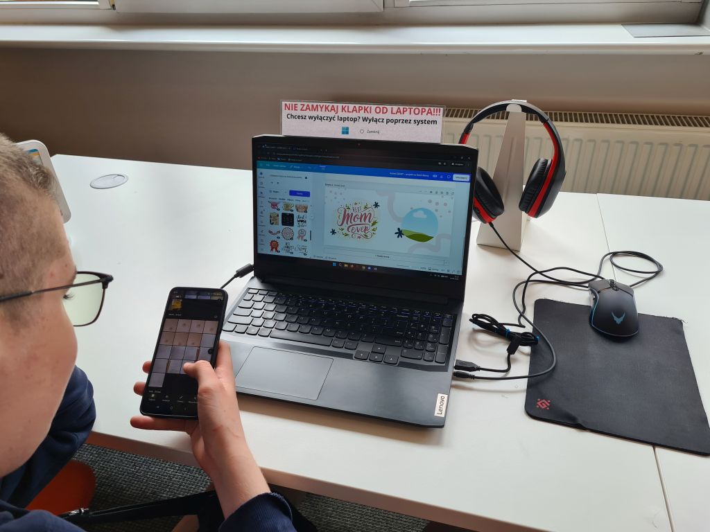 Osoba w okularach trzyma w dłoni smartfon z otwartą galerią zdjęć, siedząc przed laptopem Lenovo. Na ekranie laptopa widoczny jest projekt graficzny z napisem „Best Mom Ever”.