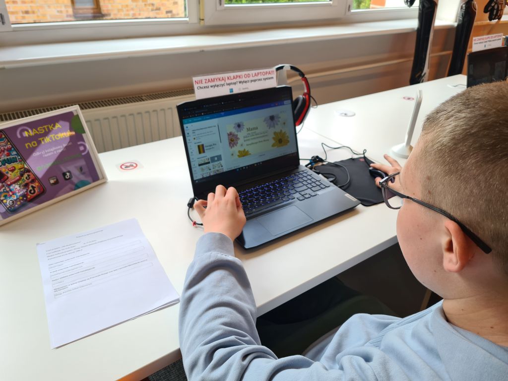 Widok z boku na osobę pracującą na laptopie, na którego ekranie widać grafikę z kwiatami i napisem „Mama”. Na białym biurku obok laptopa leży zapisana kartka papieru.