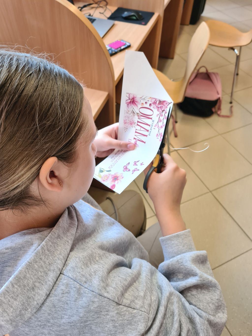 Widok z góry na osobę w szarej bluzie, która wycina nożyczkami papierowy szablon z różową grafiką i napisem „MAMO”.