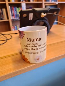 Biały kubek z nadrukiem stoi na drewnianym blacie. Na grafice widoczne są kwiaty oraz napisy zaczynające się od słów „Mama ang. (mom) Jedyna osoba...”. W tle widać rozmazany regał z książkami i tablet na podstawce.