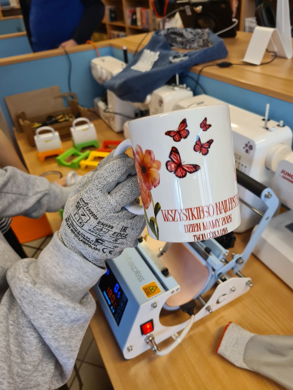 Osoba w szarej bluzie i ochronnej rękawicy roboczej trzyma gotowy biały kubek z nadrukiem przedstawiającym kwiaty, motyle oraz napis „WSZYSTKIEGO NAJLEPSZEGO DZIEŃ MAMY 2026 TWOJA CÓRKA LENA”. W tle widoczna jest maszyna do termotransferu (prasa do kubków) z wyświetlaczem oraz maszyny do szycia.