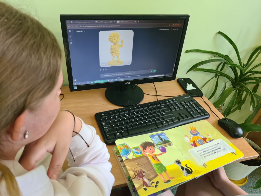 Uczestniczka warsztatów pracuje przy komputerze nad modelem postaci 3D inspirowanej książką. Obok monitora leży otwarta książka z ilustracją.