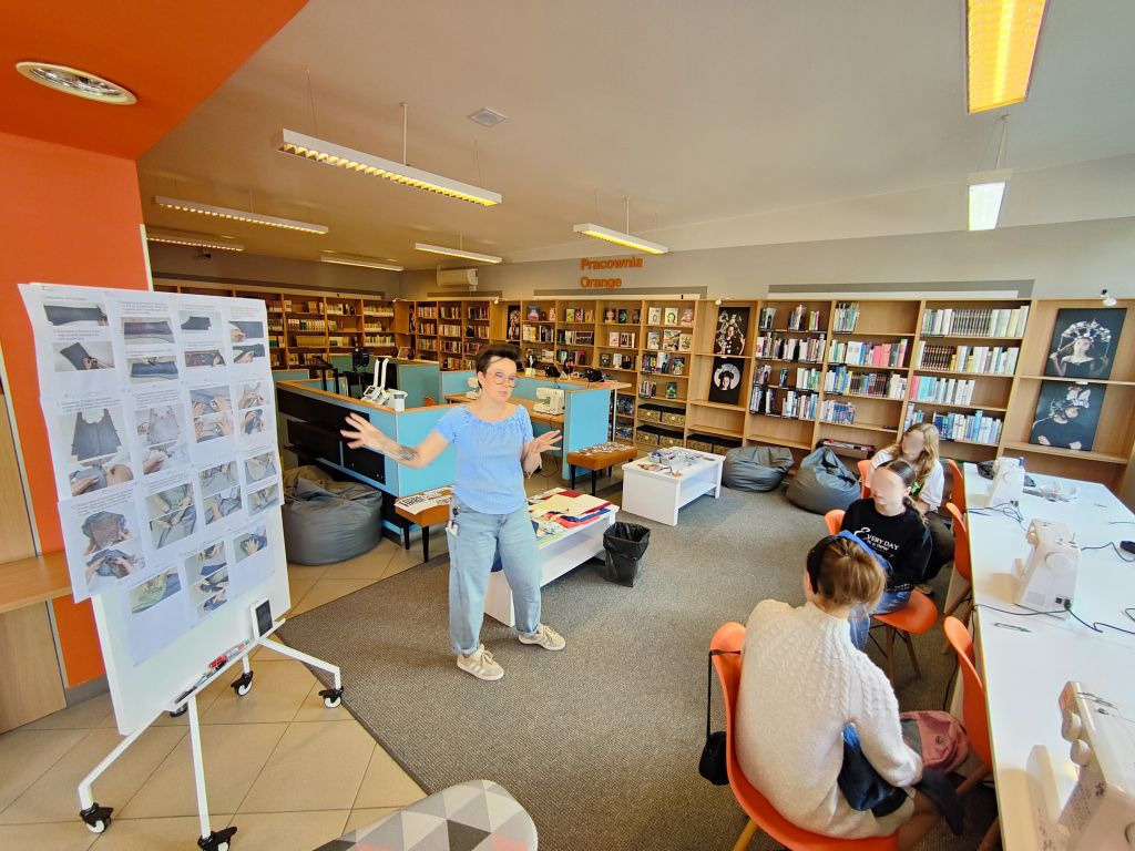 Bibliotekarka prowadzi warsztaty tworzenia toreb ze starych jeansów. Obok widoczna jest tablica z instrukcją krok po kroku, a uczestnicy siedzą przy stanowiskach.