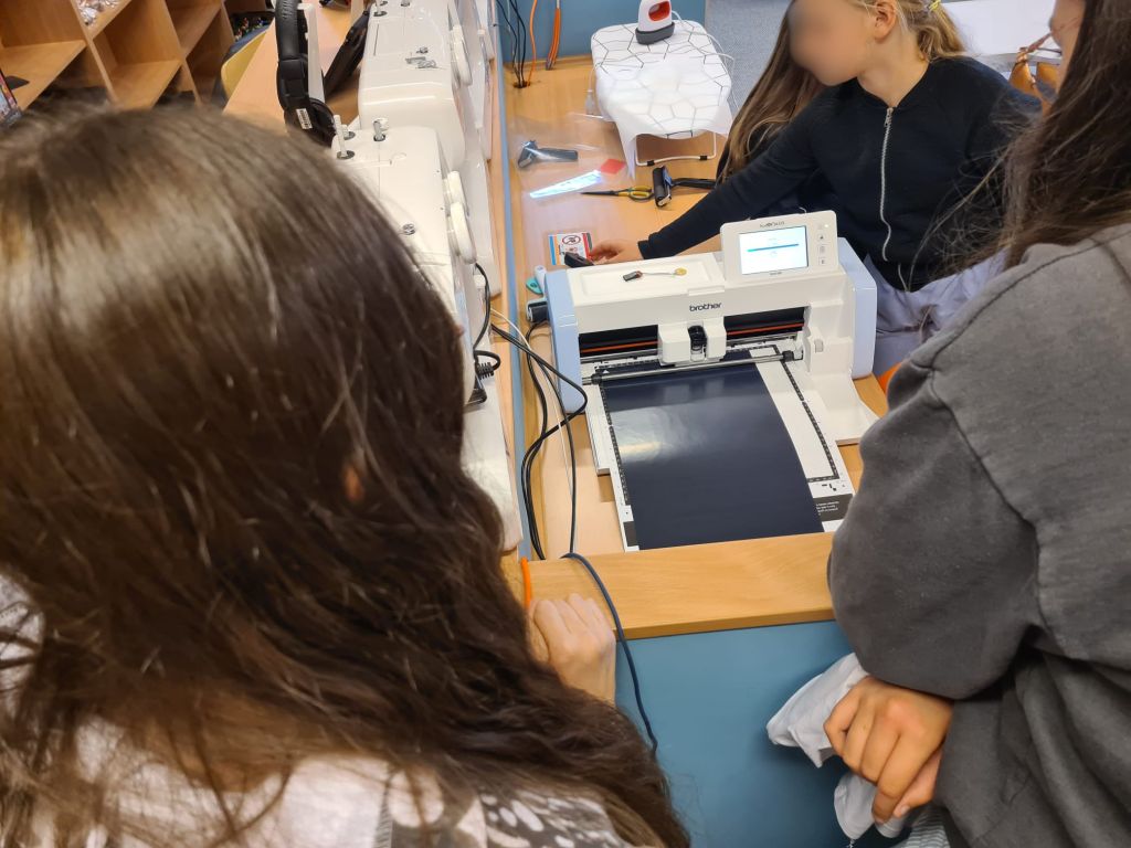 Widok z góry na stanowisko robocze, przy którym stoi kilka osób. Na niebieskim blacie umieszczony jest ploter tnący marki Brother z załadowaną ciemną folią, a obok stoją białe maszyny do szycia.