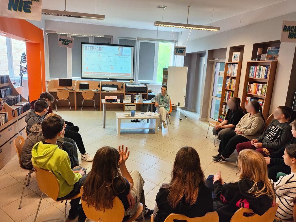 Młodzież uczestniczy w zajęciach z edukacji medialnej w Dziale dla Młodzieży NASTKA. Na ekranie wyświetlona jest prezentacja o deepfake’ach, a uczestnicy siedzą w kręgu i biorą udział w rozmowie.
