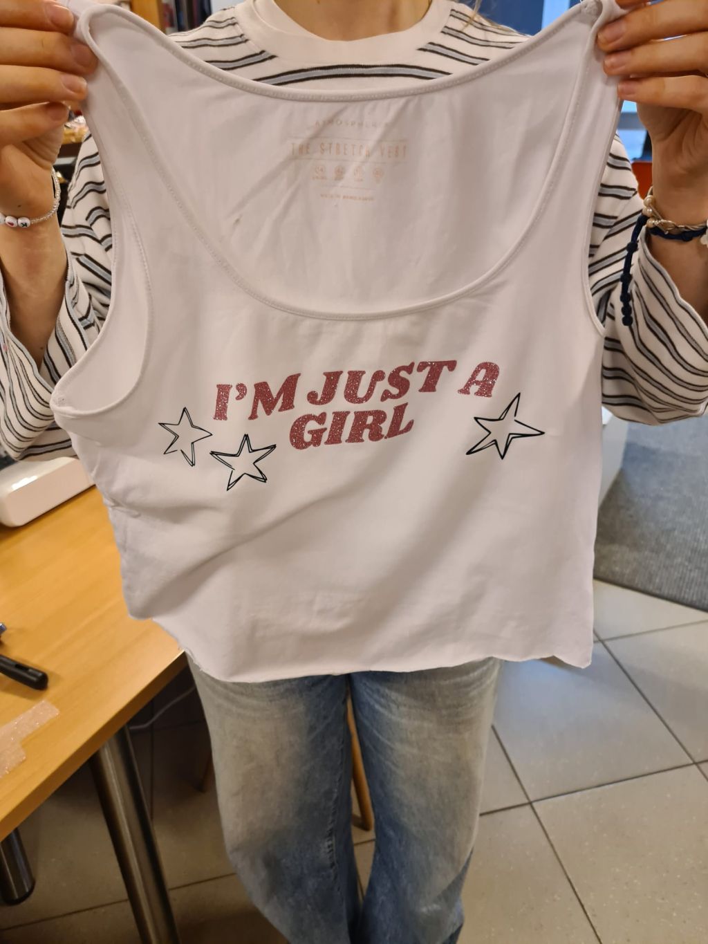 Osoba w pasiastej bluzce trzyma białą koszulkę na ramiączkach z różowym, brokatowym napisem I'M JUST A GIRL oraz trzema narysowanymi czarnym konturem gwiazdkami.
