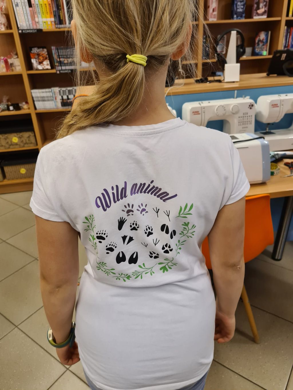 Widok od tyłu na osobę w białej koszulce z kolorowym nadrukiem. Nadruk składa się z fioletowego napisu Wild animal, okręgu z różnych śladów łap i kopyt oraz zielonych gałązek na dole.