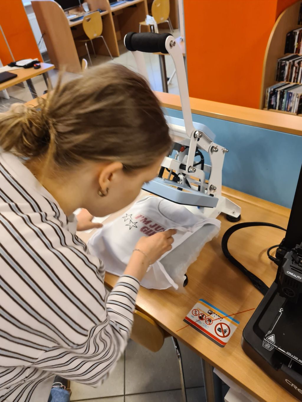 Osoba w pasiastej bluzce pochyla się nad błękitną prasą termotransferową, układając pod nią białą koszulkę z napisem I'M JUST A GIRL.