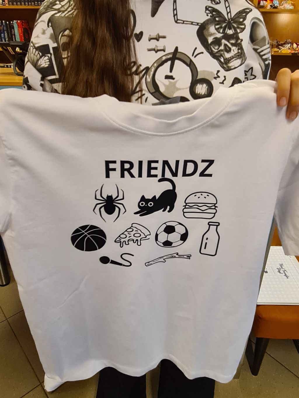 Osoba trzyma białą koszulkę z czarnym nadrukiem. Na górze widnieje napis „FRIENDZ”, a pod nim ikony: pająk, kot, hamburger, piłka do koszykówki, kawałek pizzy, piłka nożna, butelka mleka, mikrofon i patyk.