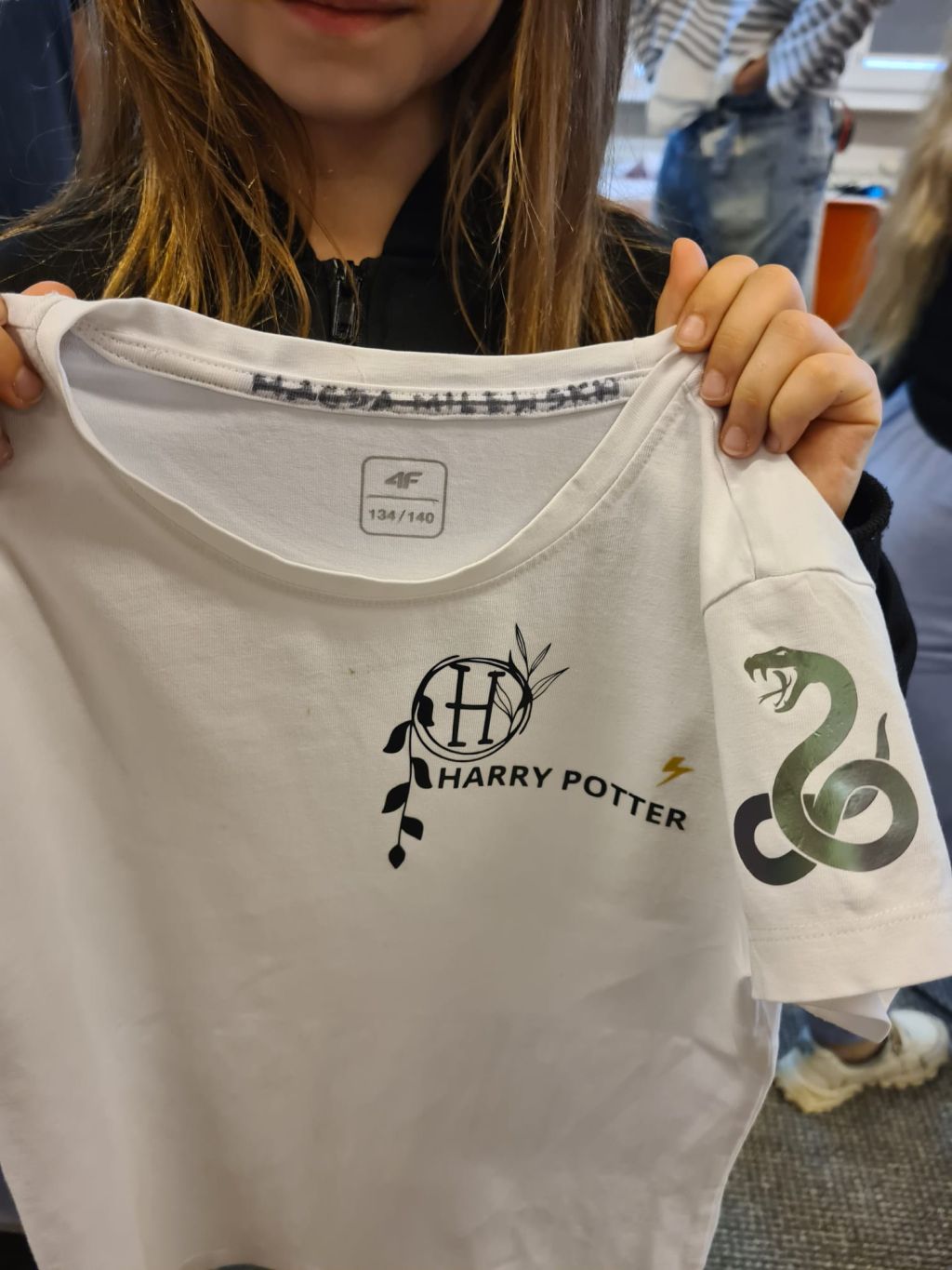 Osoba trzyma przed sobą białą koszulkę z nadrukowanym logo Harry Potter z motywem roślinnym na klatce piersiowej oraz grafiką zielonego węża na rękawie.