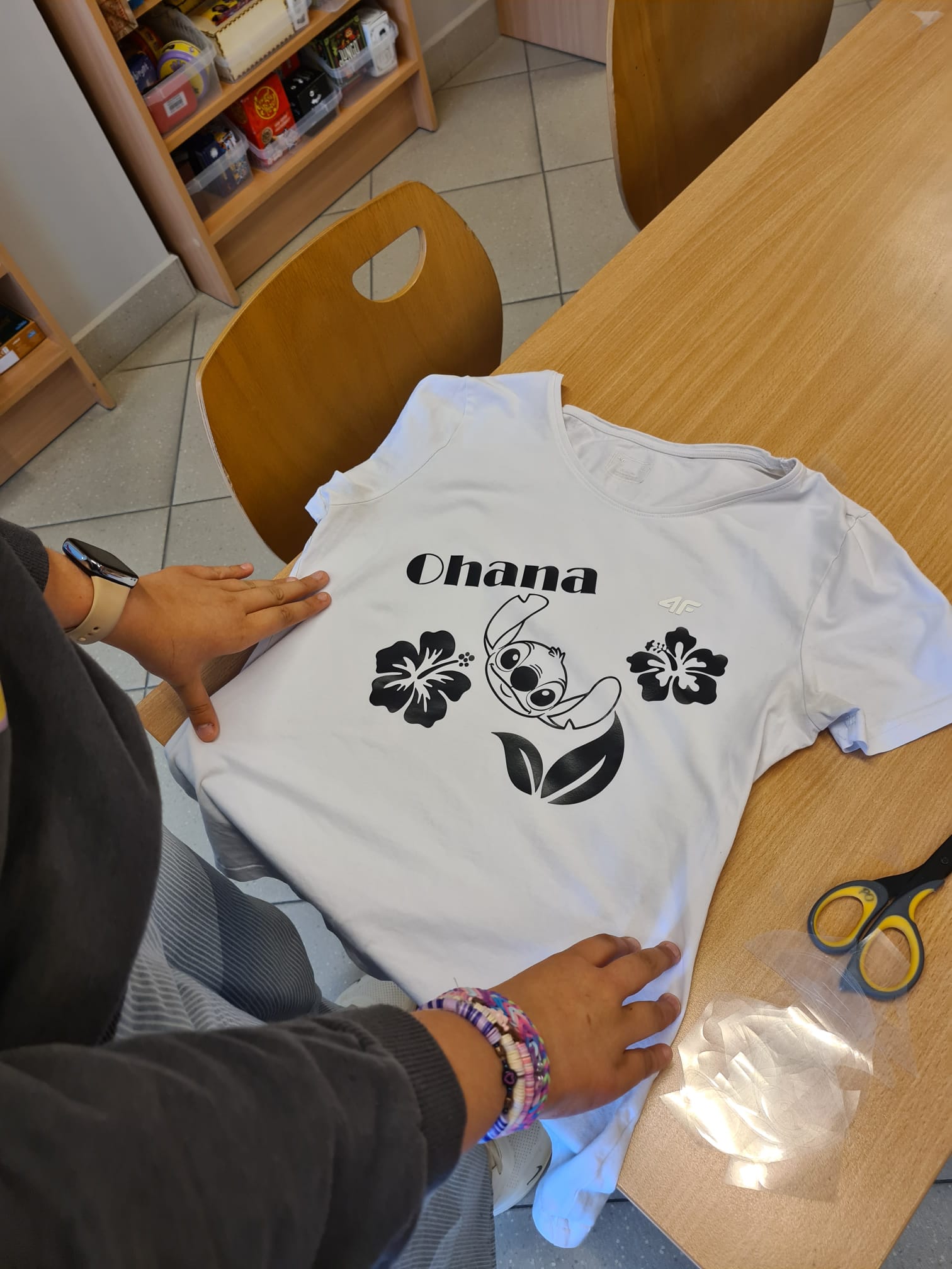 Widok z góry na dłonie wygładzające białą koszulkę leżącą na drewnianym stole. Na koszulce znajduje się czarny nadruk z postacią Stitcha, kwiatami hibiskusa i napisem Ohana. Obok leżą nożyczki i folia.