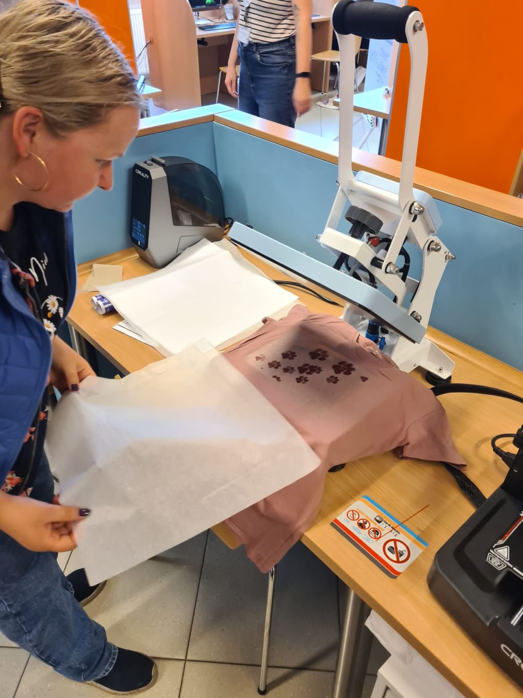 Kobieta stoi przy prasie termicznej, przygotowując różową koszulkę z nadrukiem łapek do zaprasowania, trzymając w dłoniach arkusz papieru ochronnego.