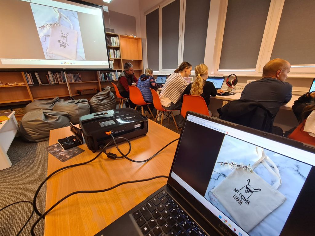 Wnętrze sali multimedialnej lub komputerowej. Kilka osób siedzi przy stanowiskach z laptopami. Na pierwszym planie widać ekran laptopa oraz duży ekran projekcyjny, na których wyświetla się zdjęcie torby płóciennej z grafiką lamy i napisem NO DRAMA LLAMA.