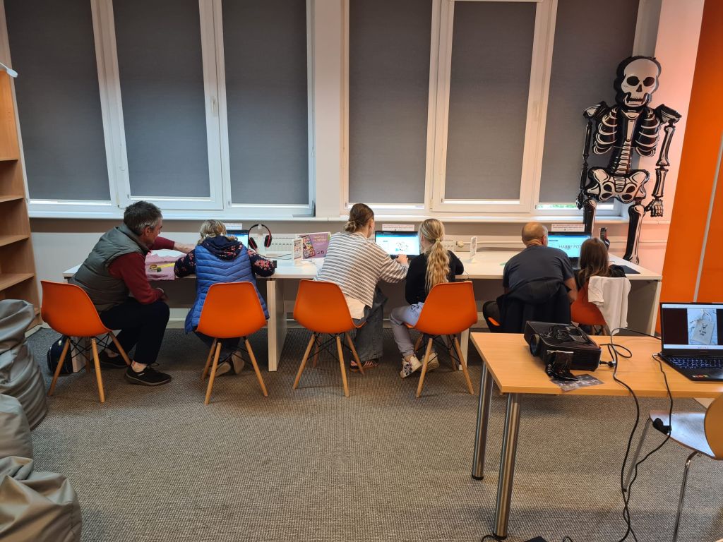 Kilka osób siedzi tyłem na pomarańczowych krzesłach przy długim białym biurku pod oknami, pracując na laptopach w jasnym pomieszczeniu. Po prawej stronie, przy ścianie, stoi dekoracyjny model szkieletu człowieka, a na pierwszym planie po prawej widać drewniany stolik z rzutnikiem i laptopem.