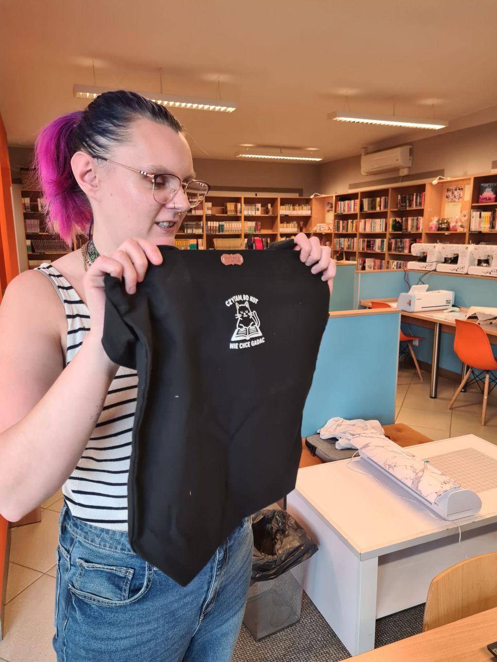 Kobieta w okularach i z fioletowo-różowymi włosami stoi w bibliotece, trzymając w dłoniach rozłożoną czarną torbę z białym nadrukiem przedstawiającym kota czytającego książkę. W tle widoczne są regały z książkami oraz maszyny do szycia.