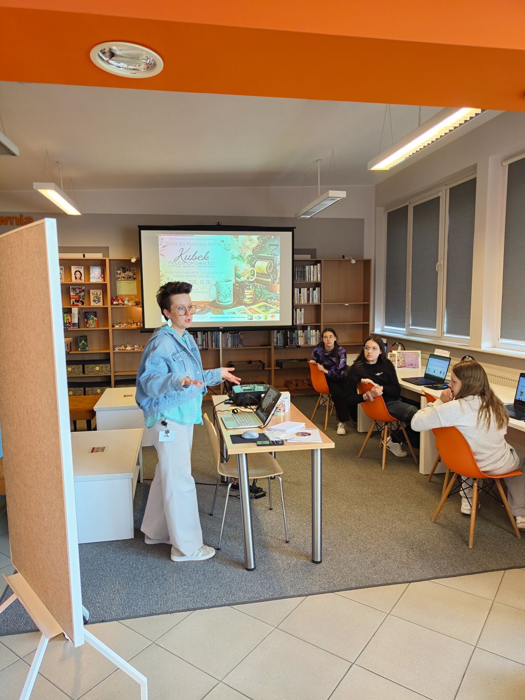 Bibliotekarka prowadzi warsztaty „Kubek pełen opowieści” w Dziale dla Młodzieży NASTKA. W tle widać ekran z plakatem wydarzenia oraz uczestników pracujących przy komputerach.