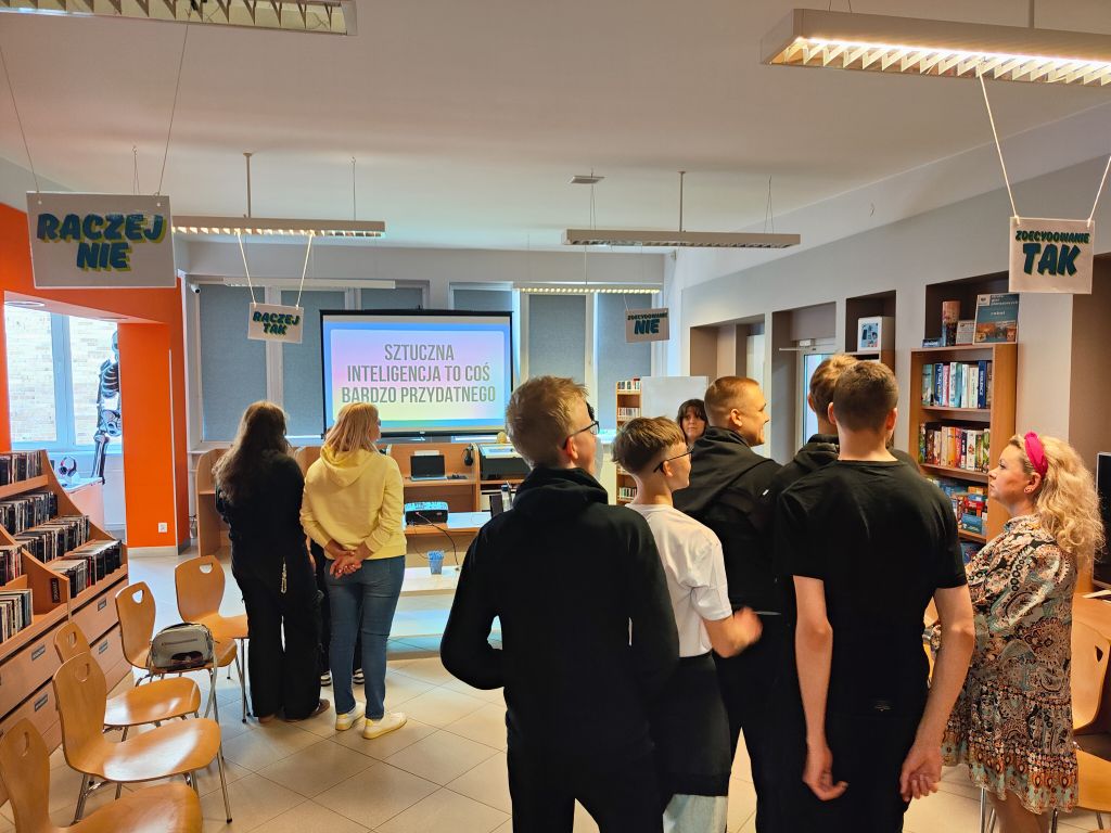 Uczestnicy stoją w sali bibliotecznej podczas ćwiczenia związanego z opiniami o sztucznej inteligencji. Na ekranie widoczny jest slajd z hasłem: „Sztuczna inteligencja to coś bardzo przydatnego”.