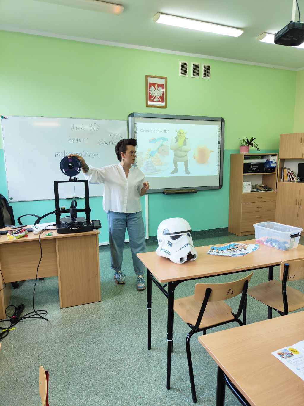 Bibliotekarka prowadzi zajęcia z druku 3D w sali lekcyjnej. Obok stoi drukarka 3D, a na ekranie wyświetlona jest prezentacja o tej technologii.