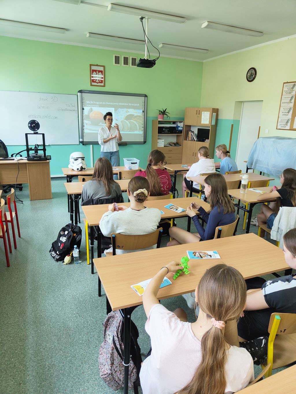 Uczniowie uczestniczą w warsztatach z druku 3D. Bibliotekarka omawia tworzenie modeli 3D, a na stolikach leżą przykładowe wydruki.
