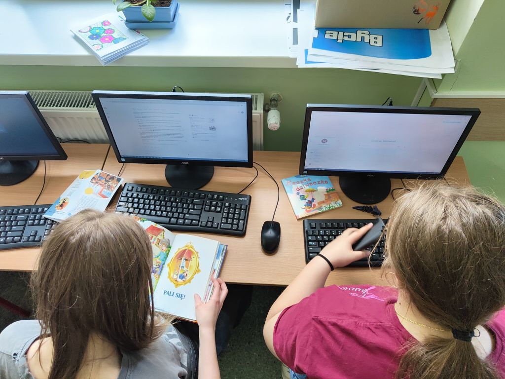 Uczennice pracują przy komputerach z książkami, przygotowując opisy postaci literackich do stworzenia modeli 3D z pomocą narzędzi AI.