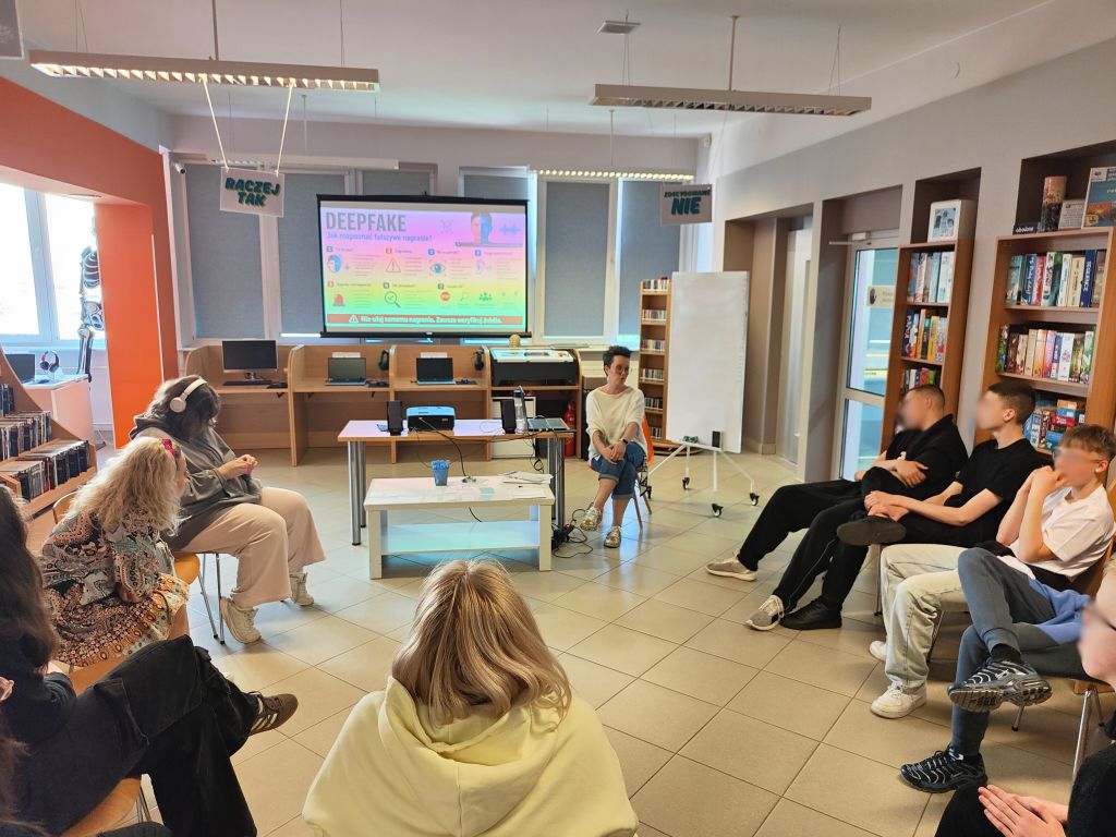 Grupa młodzieży siedzi w kręgu podczas warsztatów o edukacji medialnej. Na ekranie wyświetlona jest prezentacja „Deepfake. Jak rozpoznać fałszywe nagranie?”.