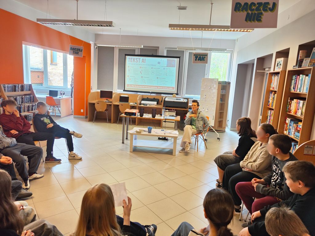 Bibliotekarka prowadzi zajęcia o rozpoznawaniu tekstów tworzonych przez sztuczną inteligencję. Uczestnicy siedzą w kręgu, a na ekranie widoczna jest prezentacja „Tekst AI”.