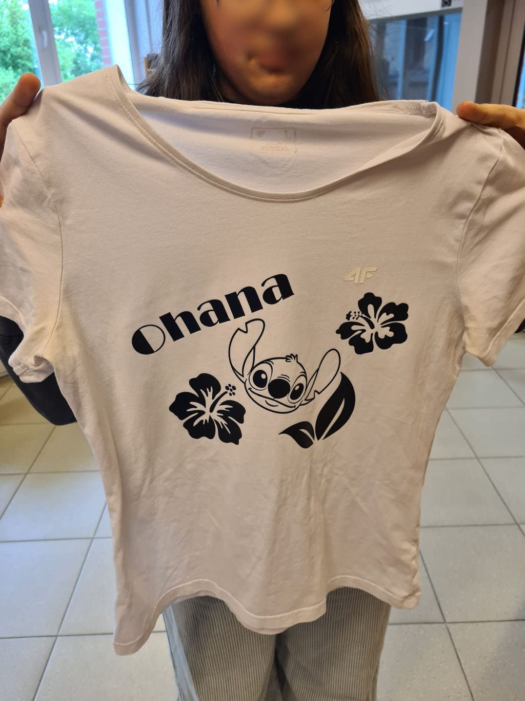 Osoba trzyma przed sobą białą koszulkę z czarnym nadrukiem przedstawiającym postać Stitcha, dwa kwiaty hibiskusa oraz ukośny napis Ohana.