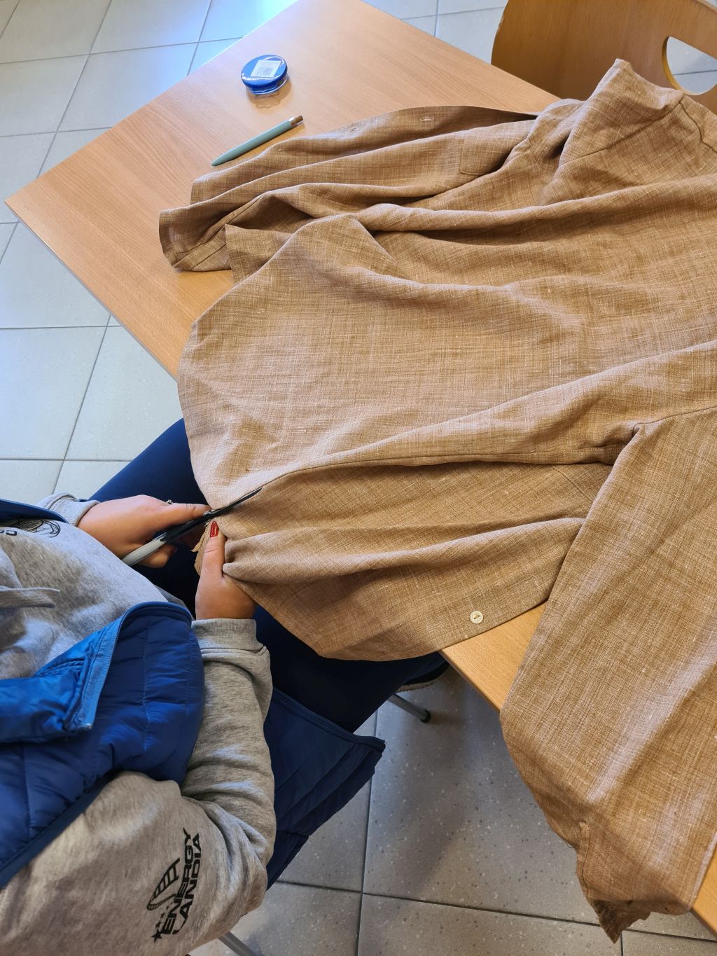 Widok z góry na osobę w szarej bluzie i niebieskiej kamizelce, która rozcina nożyczkami brązową, lnianą koszulę ułożoną na drewnianym stole.
