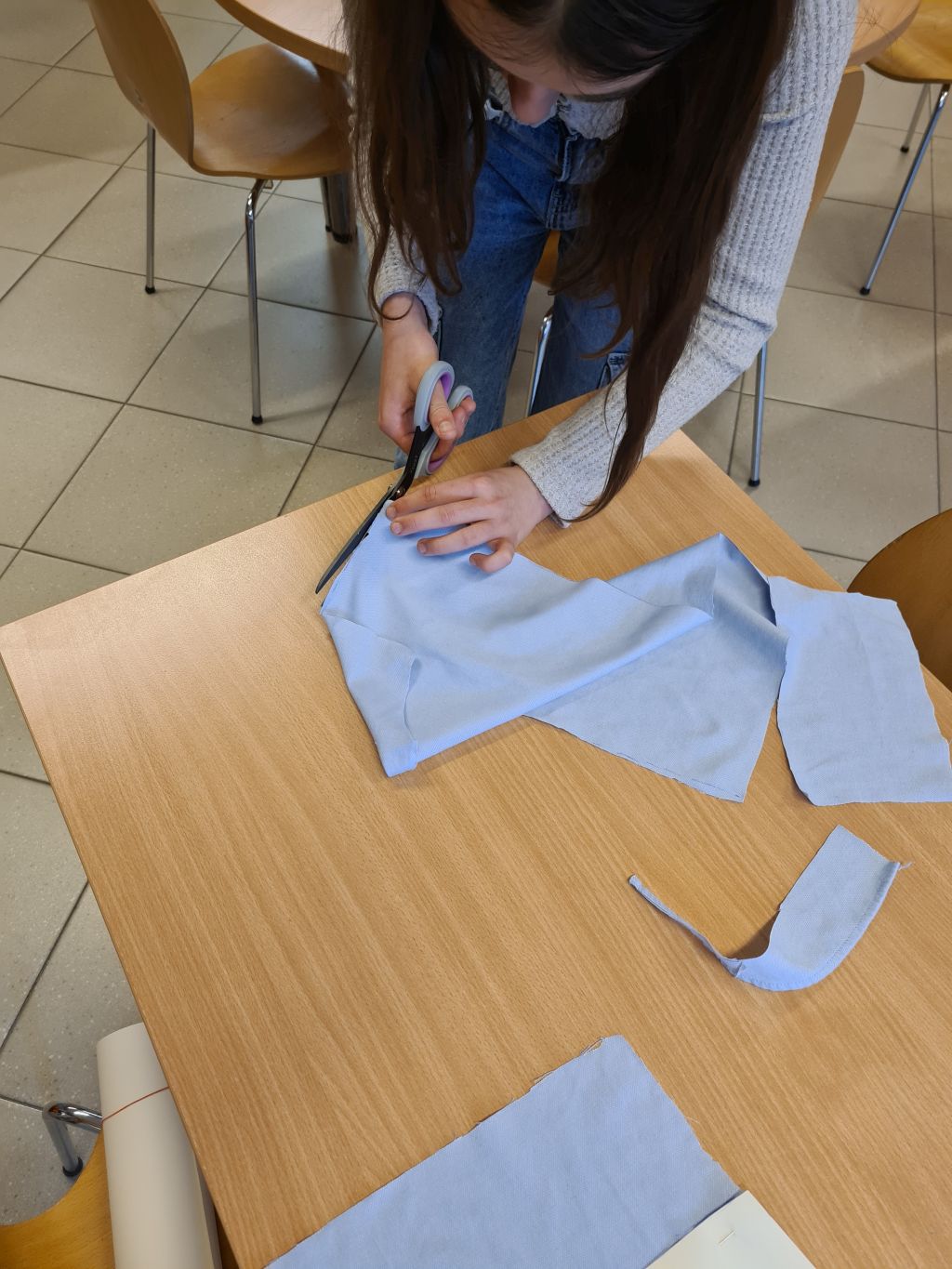Osoba w jasnym swetrze i jeansach stoi przy drewnianym stole i wycina nożyczkami kształty z błękitnego materiału.