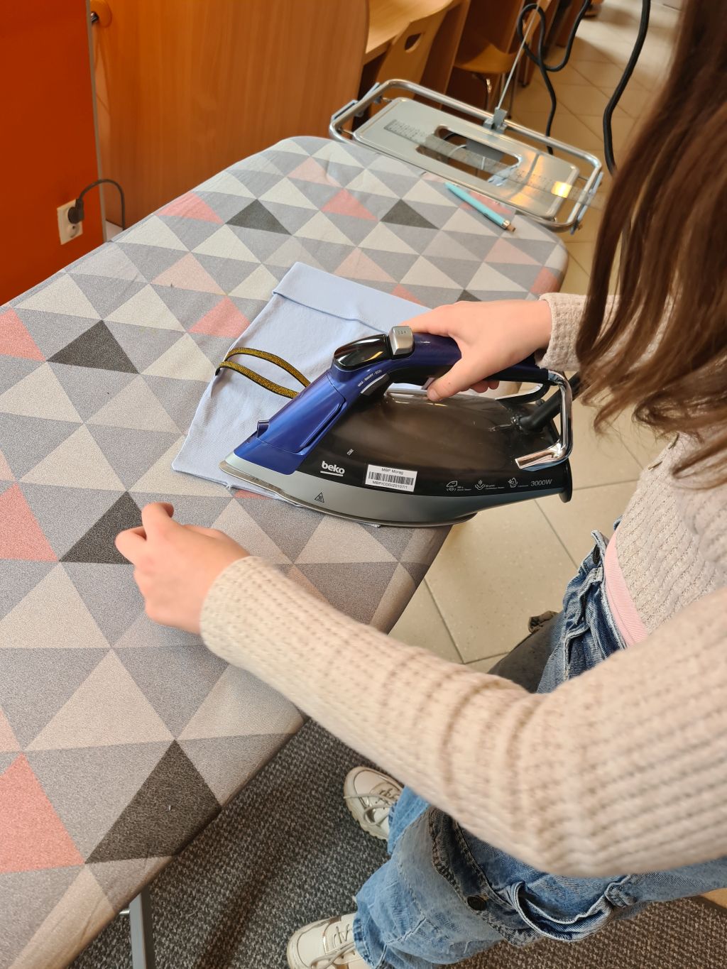 Zbliżenie z góry na osobę ubraną w beżowy sweter i dżinsy, prasującą jasnoniebieski materiał na desce do prasowania. W tle rozmyty widok na inne stoliki i krzesła w bibliotece.