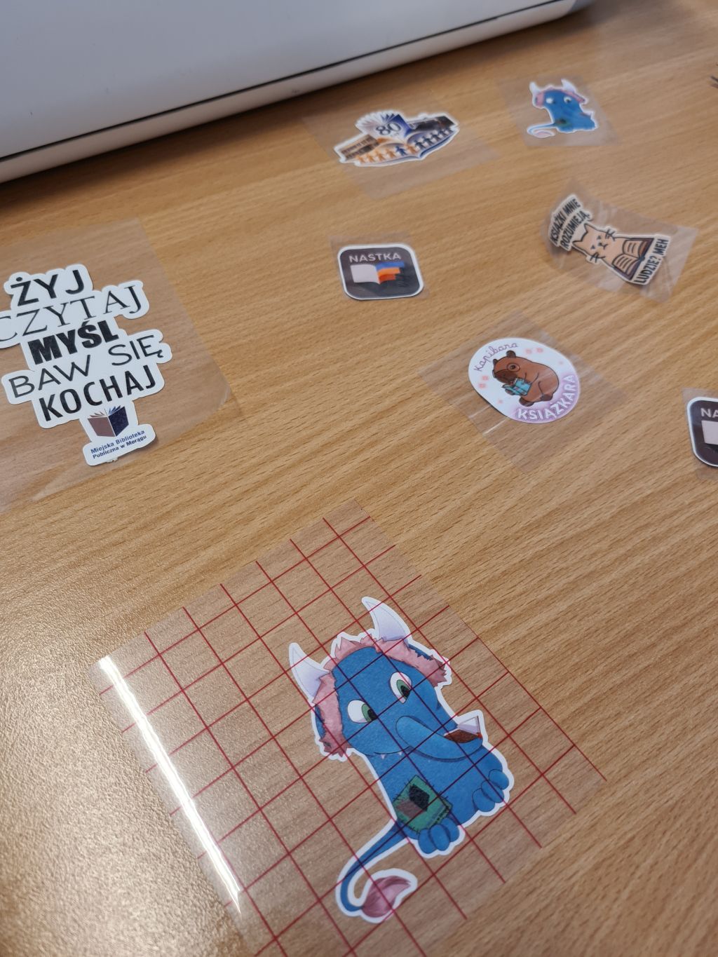 Zbliżenie na drewniany blat, na którym leżą przezroczyste naklejki termotransferowe z grafikami (m.in. niebieski stworek, kapibara) i napisami, w tym głównym: „ŻYJ CZYTAJ MYŚL BAW SIĘ KOCHAJ”.