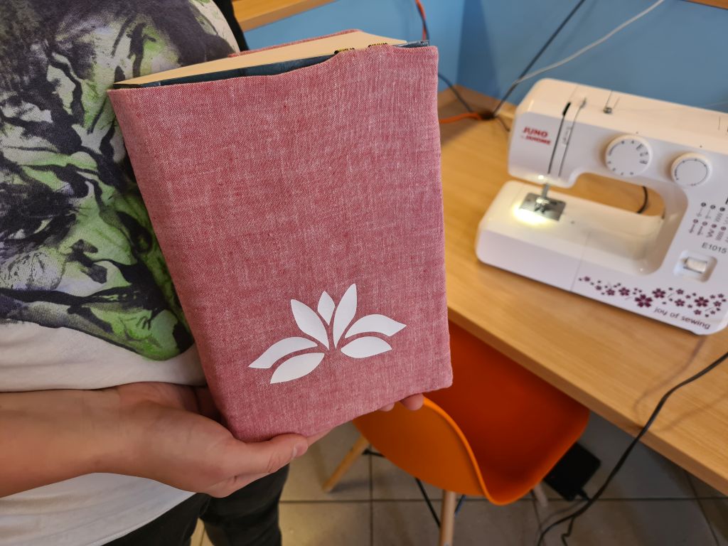 Osoba trzyma książkę ubraną w różowo-czerwony, lniany otulacz z naklejoną białą grafiką przedstawiającą kwiat lotosu. W tle, na biurku, stoi biała maszyna do szycia i pomarańczowe krzesło.