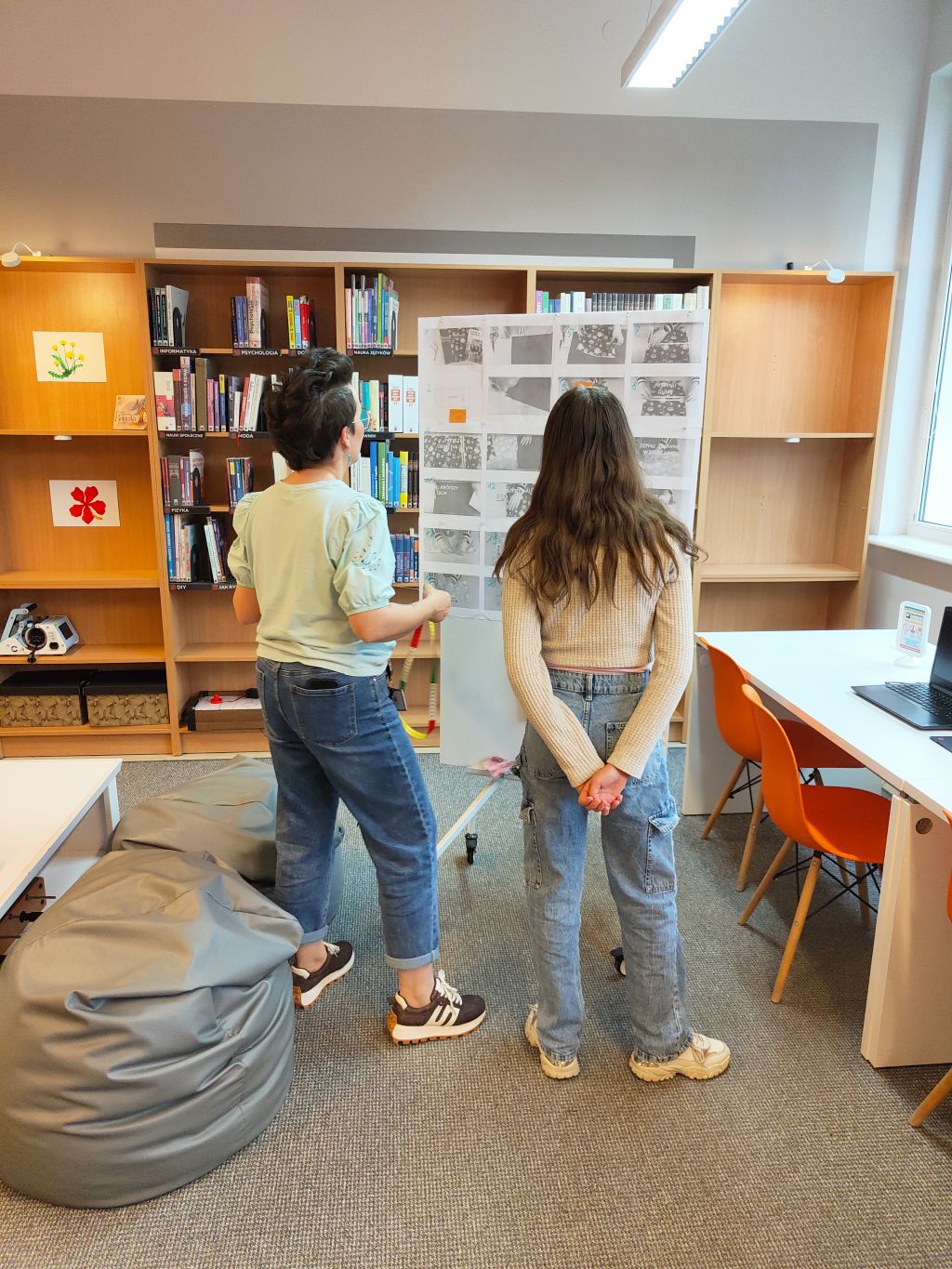 Dwie osoby stoją tyłem w pomieszczeniu bibliotecznym i oglądają białą tablicę (flipchart) z przypiętymi czarno-białymi instrukcjami obrazkowymi. W tle widać regały pełne książek, pufy oraz biurko z laptopem.