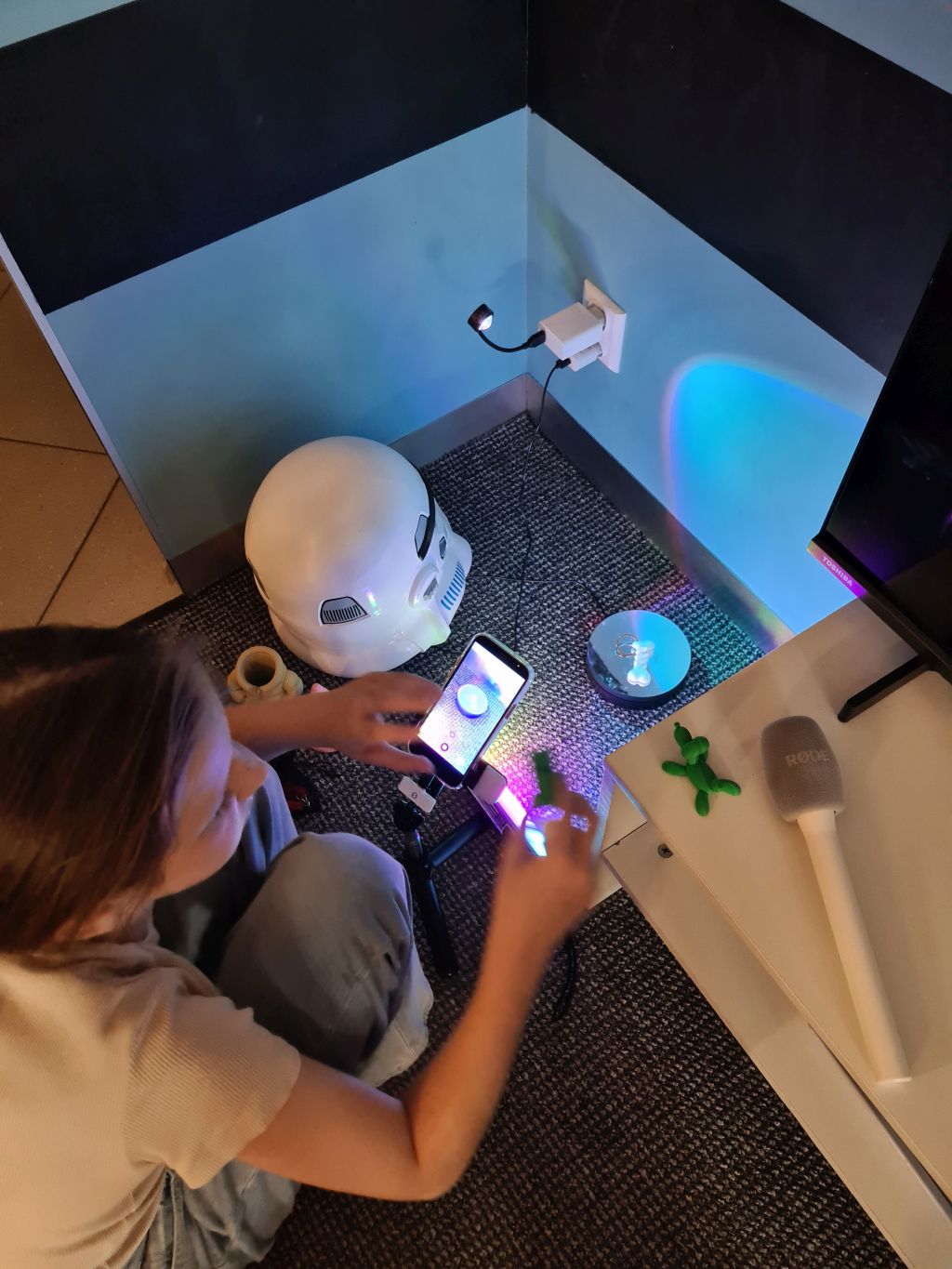 Dziewczyna siedząca na podłodze, ustawiająca dłońmi smartfon na małym statywie. W tle na podłodze leży hełm Szturmowca, a obok na stoliku stoi mikrofon i mała zielona figurka.