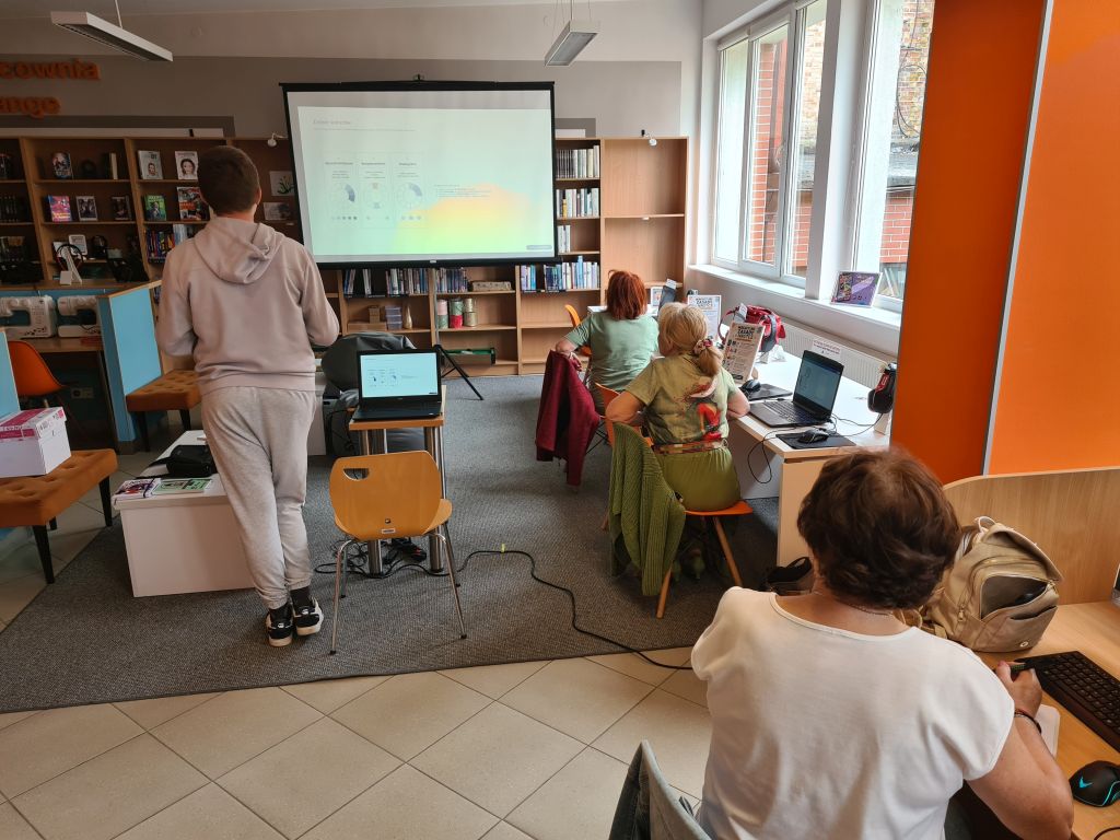 Uczestniczki warsztatów komputerowych siedzą przy laptopach w Dziale dla Młodzieży NASTKA. Prowadzący stoi obok ekranu projekcyjnego i omawia prezentowane zagadnienia.