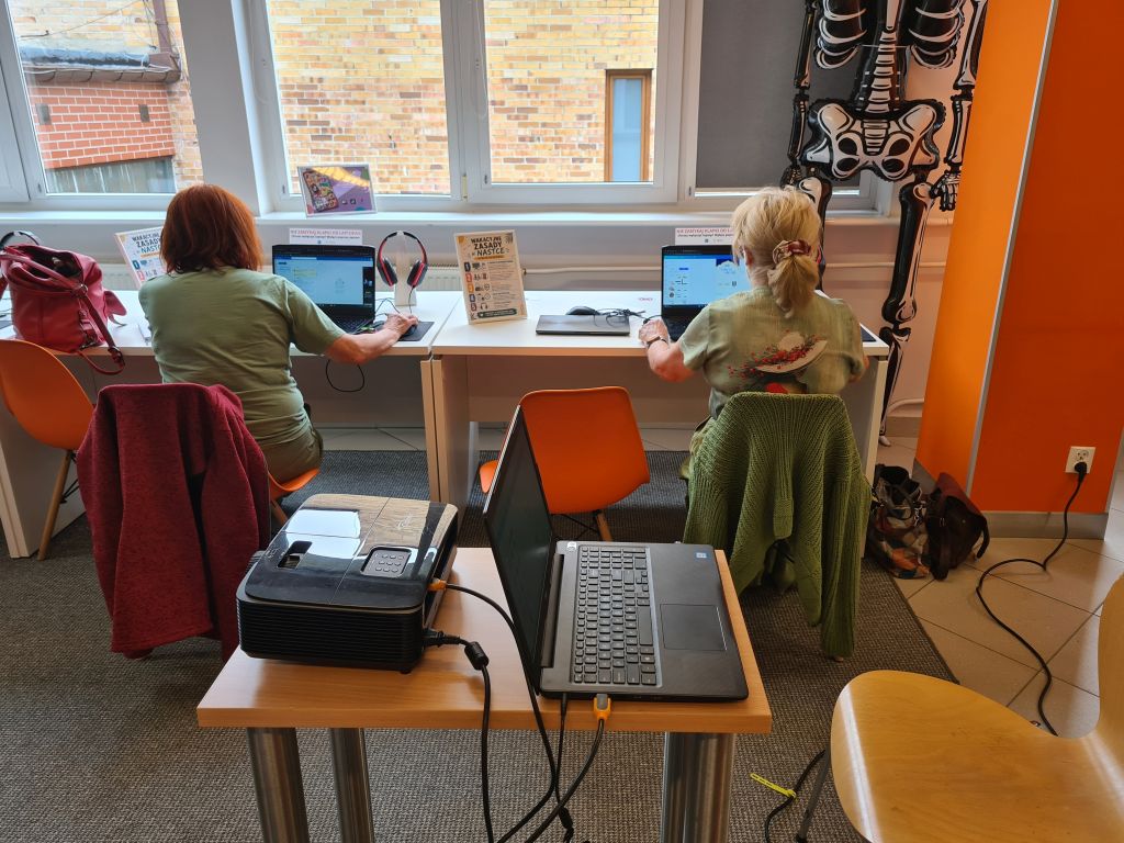 Dwie uczestniczki pracują przy laptopach ustawionych pod oknem. Na pierwszym planie znajdują się projektor oraz komputer prowadzącego.